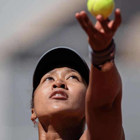 Eine asiatisch aussehende Frau schaut bei Sonnenschein hoch zum Tennisball, den ihre linke Hand gerade hochwirft