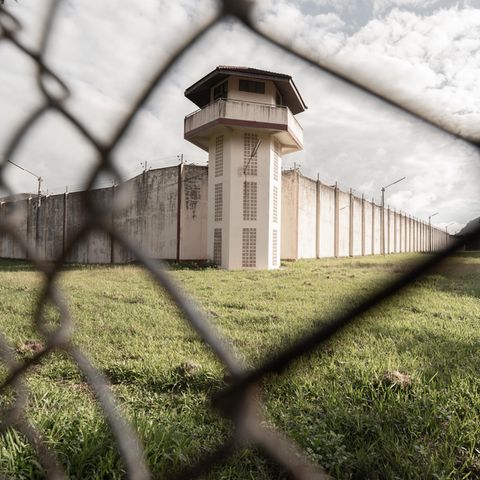Blick durch Maschendrahtzaun auf Gefängnismauer und Wachturm
