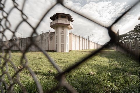 Blick durch Maschendrahtzaun auf Gefängnismauer und Wachturm