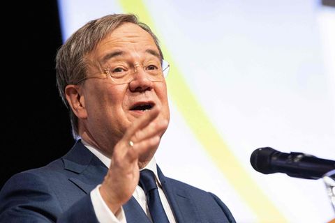 Ein weißer Mann in blauem Anzug steht vor einem Mikrofon und gestikuliert mit seiner rechten Hand auf Höhe seines Kinns