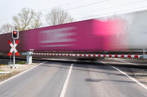Hinter zwei geschlossenen Schranken quert ein Güterzug mit Containern eine Straße. Die Waggons sind wegen des Tempos unscharf