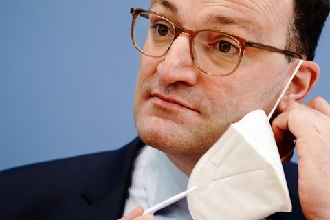 Ein weißer Mann mit braunen Locken und dünner Hornbrille nimmt eine FFP2-Maske ab, die mit einem Gummi noch am linken Ohr hängt