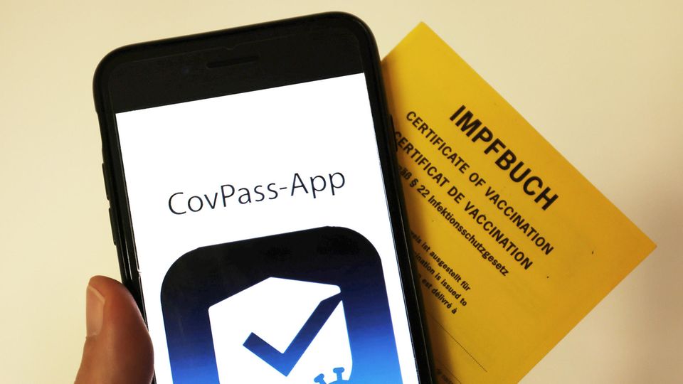 Eine Hand hält einen gelben Impfpass und ein Smartphone mit der "CovPass-App" auf dem Display hoch