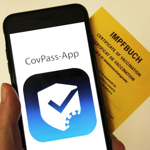 Eine Hand hält einen gelben Impfpass und ein Smartphone mit der "CovPass-App" auf dem Display hoch