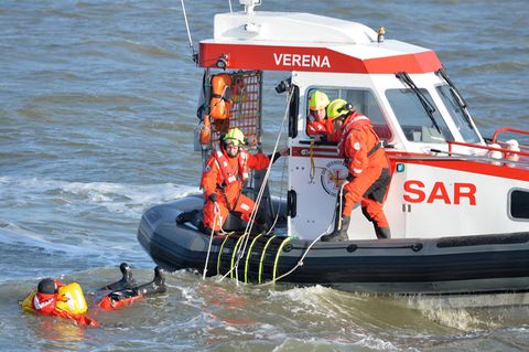 Zwei Personen in rot-schwarzen Overalls und gelben Helmen stehen am Rand eines Boots und schauen auf einen im Wasser treibenden
