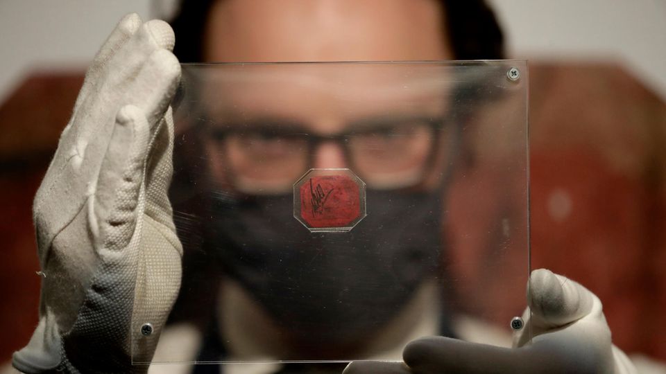 Ein weißer Mann hält sich mit weißen Handschuhen eine Plexiglasscheibe mit einer magenta-farbenen Briefmarke vors Gesicht
