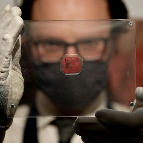 Ein weißer Mann hält sich mit weißen Handschuhen eine Plexiglasscheibe mit einer magenta-farbenen Briefmarke vors Gesicht