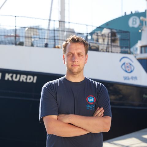 Ein Mann steht mit verschränkten Armen vor einem Schiff mit dem Namen Alan Kurdi