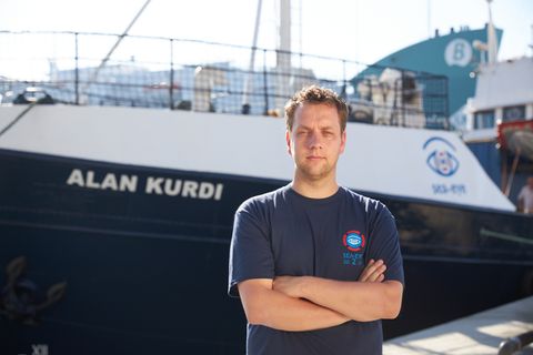 Ein Mann steht mit verschränkten Armen vor einem Schiff mit dem Namen Alan Kurdi