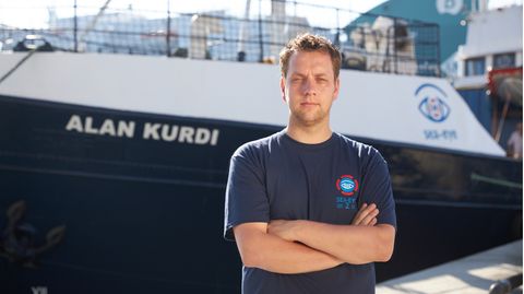 Ein Mann steht mit verschränkten Armen vor einem Schiff mit dem Namen Alan Kurdi