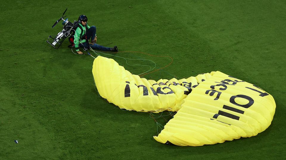 Ein Greenpeace-Aktivist sitzt mit einem gelben Fallschirm auf dem Fußballrasen in München