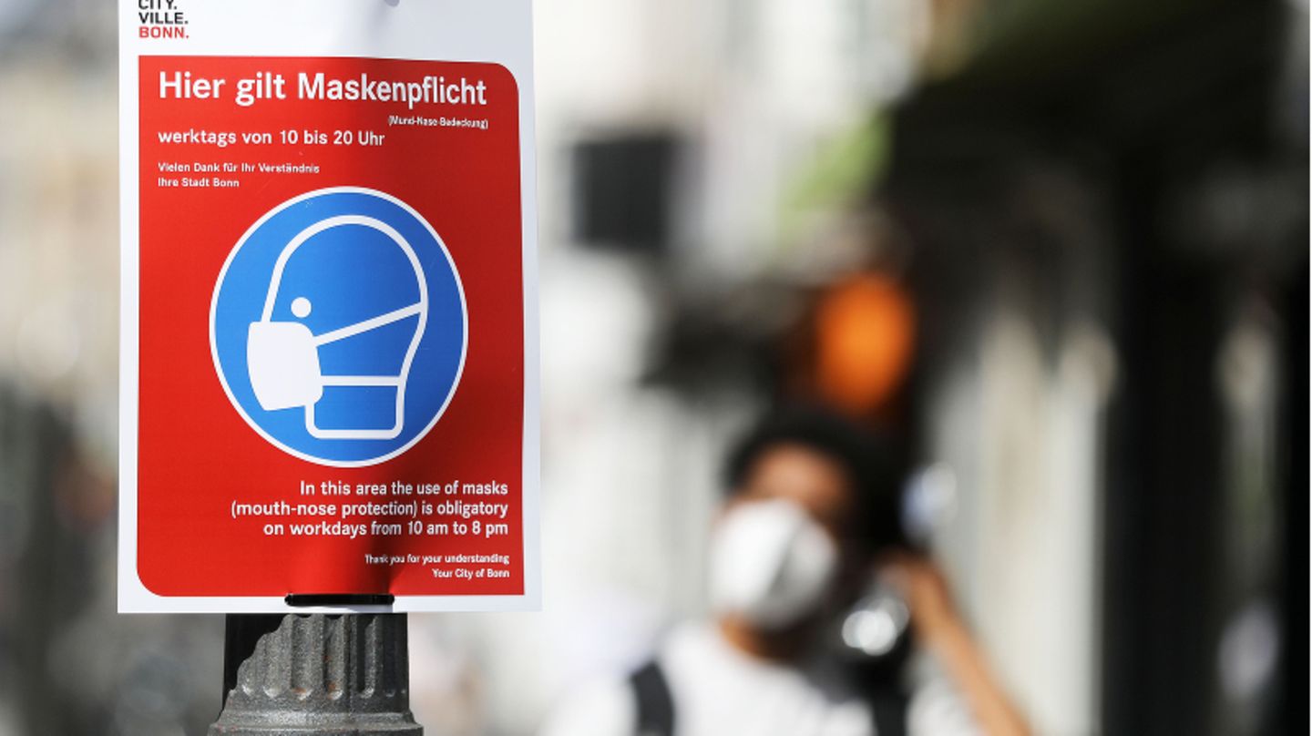 Ein Schild weist auf die Maskenpflicht hin, im Hintergrund sieht man verschwommen eine Person mit Mund-Nasen-Schutz