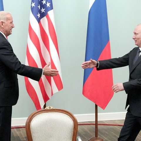 Joe Biden und Wladimir Putin reichen sich die Hände vor den Flaggen von Russland und den USA