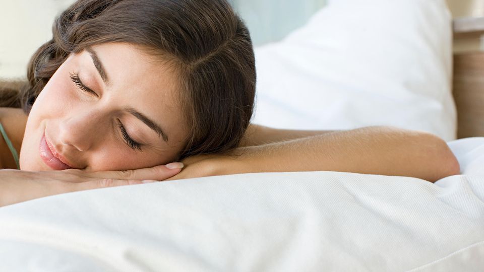 Eine junge weiße Frau mit langen, braunen Haaren liegt mit geschlossenen Augen und leicht lächelnd in einem weiß bezogenen Bett