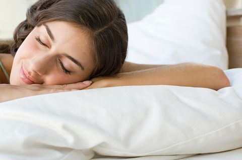 Eine junge weiße Frau mit langen, braunen Haaren liegt mit geschlossenen Augen und leicht lächelnd in einem weiß bezogenen Bett