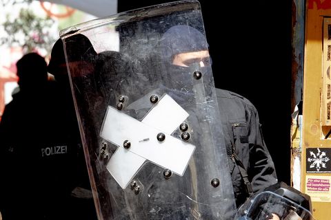 Ein Polizist mit Sturmhaube verlässt hinter einem Schild und mit Helm in der linken Hand ein heruntergekommenes Haus