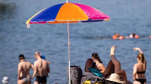 An einem Strand steht ein bunter Sonnenschirm. Erwachsene stehen in Badekleidung im Sand und schauen aufs Wasser