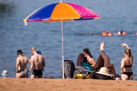 An einem Strand steht ein bunter Sonnenschirm. Erwachsene stehen in Badekleidung im Sand und schauen aufs Wasser