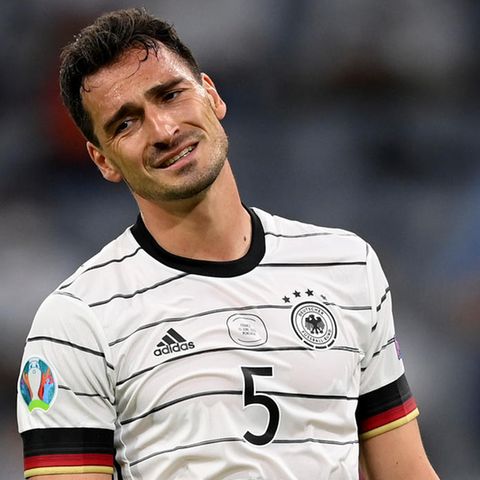 Ein weißer Mann mit kurzen, dunkelbraunen Locken steht im weißen DFB-Trikot im Stadion und lchelt gequält