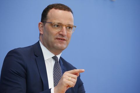 Ein Mann mit kurzen braunen Locken sitzt in Anzug vor einer blauen Wand und weist mit dem rechten Zeigefinger von sich