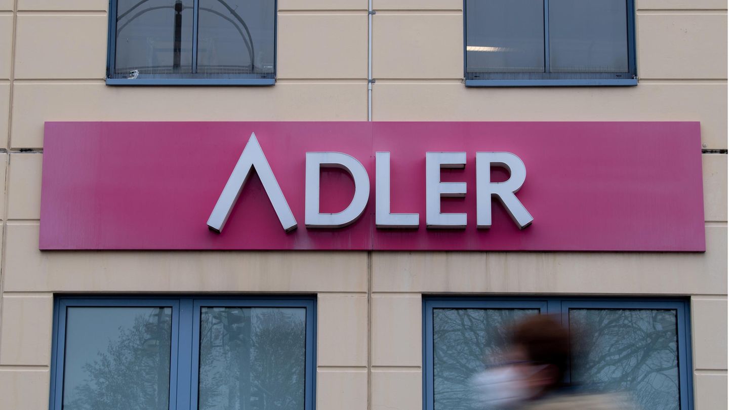 Eine Frau mit Mund-Nase-Bedeckung geht an einem Schild des Unternehmens Adler Modemärkte an einem Einkaufszentrum vorbei