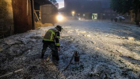 Ein Feuerwehrmann steht in einem Reiterhof in Hagelkörnern und Wasser und stellt eine Pumpe auf