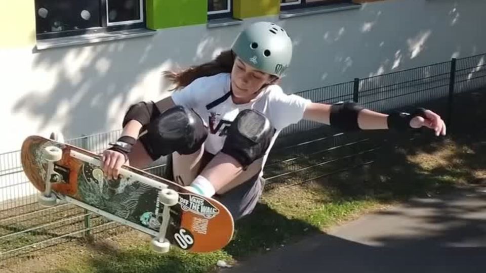Eine junge weiße Teenagerin mit Helm und Ellenbogen- und Knieschonern springt mit einer Hand am Skateboard aus einer Halfpipe