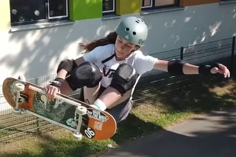 Eine junge weiße Teenagerin mit Helm und Ellenbogen- und Knieschonern springt mit einer Hand am Skateboard aus einer Halfpipe