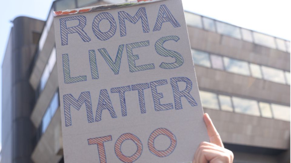 Ein Mann hält ein Pappschild hoch, auf dem Roma Lives Matter Too steht