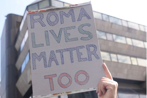 Ein Mann hält ein Pappschild hoch, auf dem Roma Lives Matter Too steht