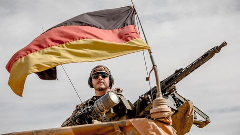 Ein Bundeswehr-Soldat schaut mit Sonnenbrille und schwarzen Ohrenschützern unter einer deutschen Flagge von einem Fahrzeug herab
