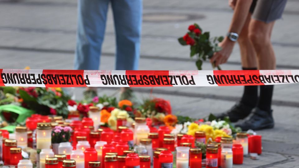 Unter einem Flatterband mit "Polizeiabsperrung"-Aufdruck stehen Kerzen und liegen Blumen, zwei Menschen stehen im Hintergrund