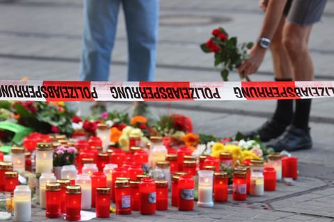 Unter einem Flatterband mit "Polizeiabsperrung"-Aufdruck stehen Kerzen und liegen Blumen, zwei Menschen stehen im Hintergrund