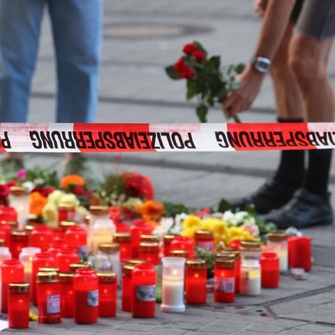 Unter einem Flatterband mit "Polizeiabsperrung"-Aufdruck stehen Kerzen und liegen Blumen, zwei Menschen stehen im Hintergrund