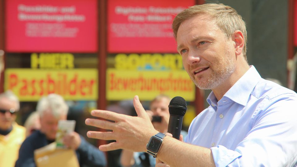 Christian Lindner mit Mikrofon in Erklärerpose vor rot-gelben Aufklebern