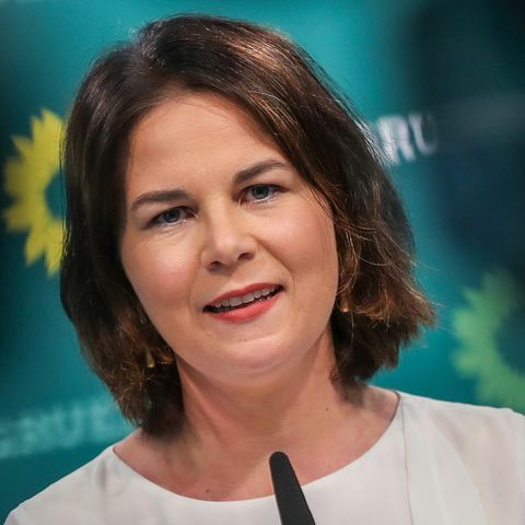 Eine weiße Frau mit dunkelbraunen, schulterlangen Haaren steht an einem Mikrofon or einer grünen Wand mit Sonnenblumen darauf