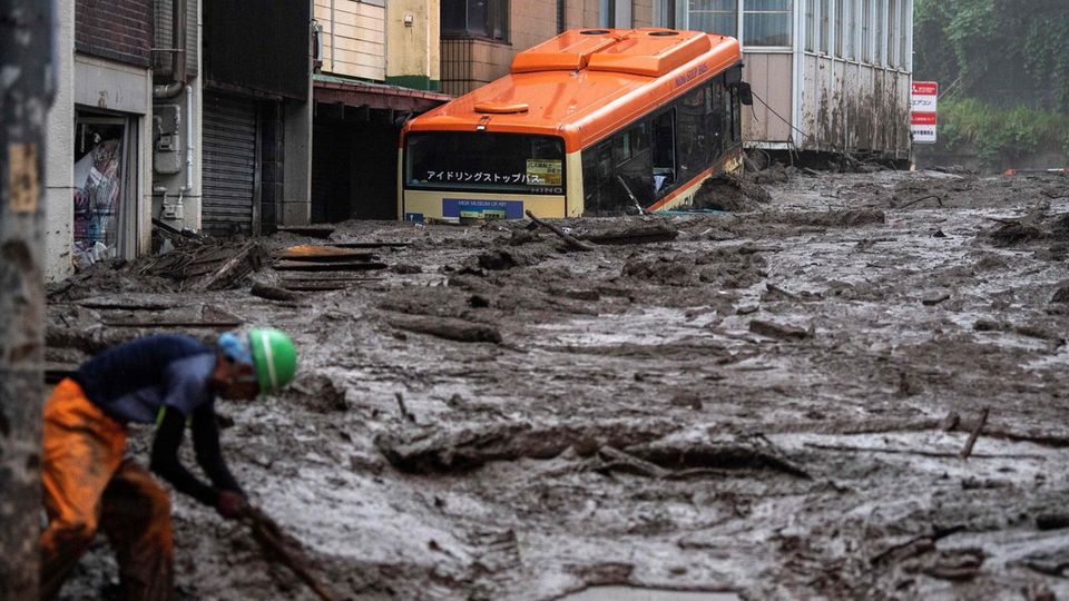 Japan Bus Schlammlawine Überschwemmungen Ein Armeiter in oranger Gummihose und grünem Sicherheitshelm schaufelt Schlamm, während im Hintergrund ein oranger Bus steckt