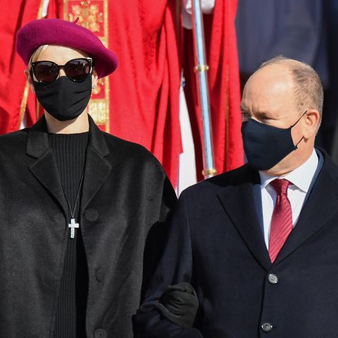 Eine elegant gekleidete Frau mit weinroter Baskenmütze, Sonnenbrille und schwarzem Mundschutz steht neben einem kleineren Mann