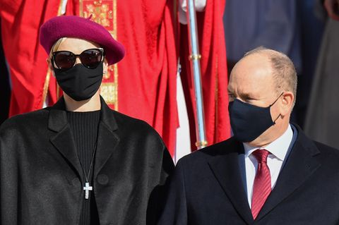 Eine elegant gekleidete Frau mit weinroter Baskenmütze, Sonnenbrille und schwarzem Mundschutz steht neben einem kleineren Mann