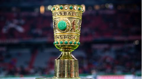Auf einem Sockel in einem Fußball-Stadion steht er goldene DFB-Pokal mit grünen Ziersteinen