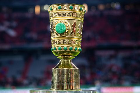 Auf einem Sockel in einem Fußball-Stadion steht er goldene DFB-Pokal mit grünen Ziersteinen