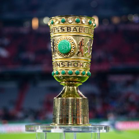 Auf einem Sockel in einem Fußball-Stadion steht er goldene DFB-Pokal mit grünen Ziersteinen