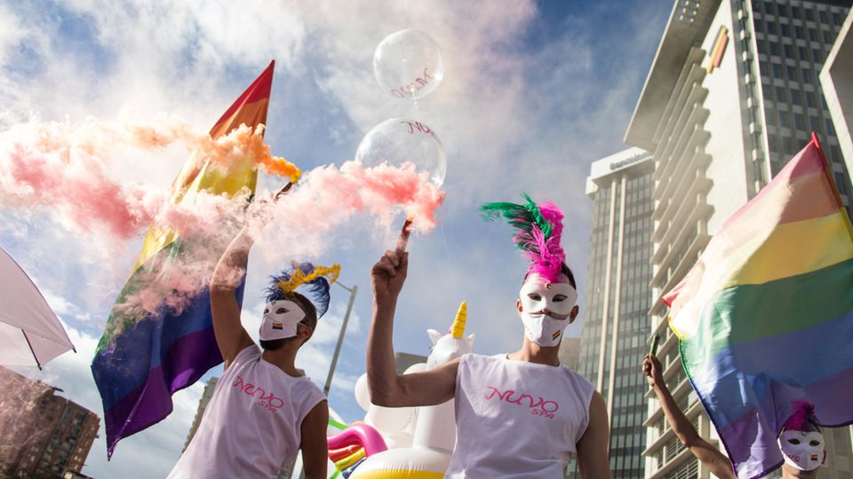 En un desfile del orgullo, la gente camina con ropa colorida con máscaras y agita la bandera del arco iris.