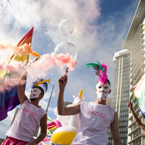 Bei einer Pride-Parade marschieren bunt gekleidete Menschen mit Masken und schwenken die Regenbogen-Flagge