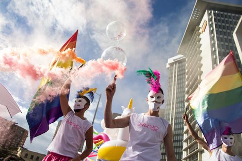 Bei einer Pride-Parade marschieren bunt gekleidete Menschen mit Masken und schwenken die Regenbogen-Flagge