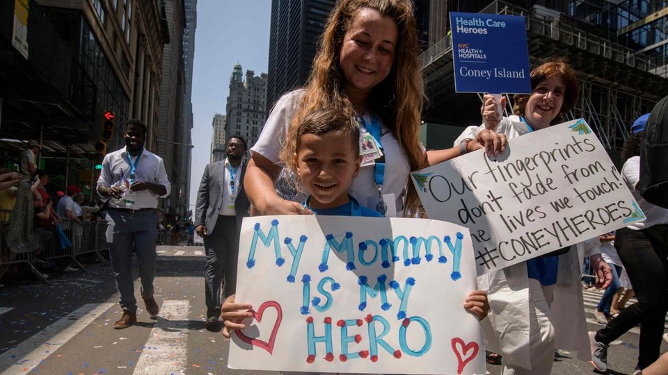 Ein blonder weißer Junge hält ein Schild, auf dem steht: "Meine Mama ist meine Heldin". Von hinten umarmt ihr eine Frau