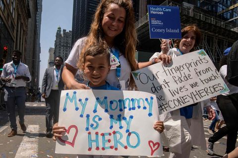 Ein blonder weißer Junge hält ein Schild, auf dem steht: "Meine Mama ist meine Heldin". Von hinten umarmt ihr eine Frau