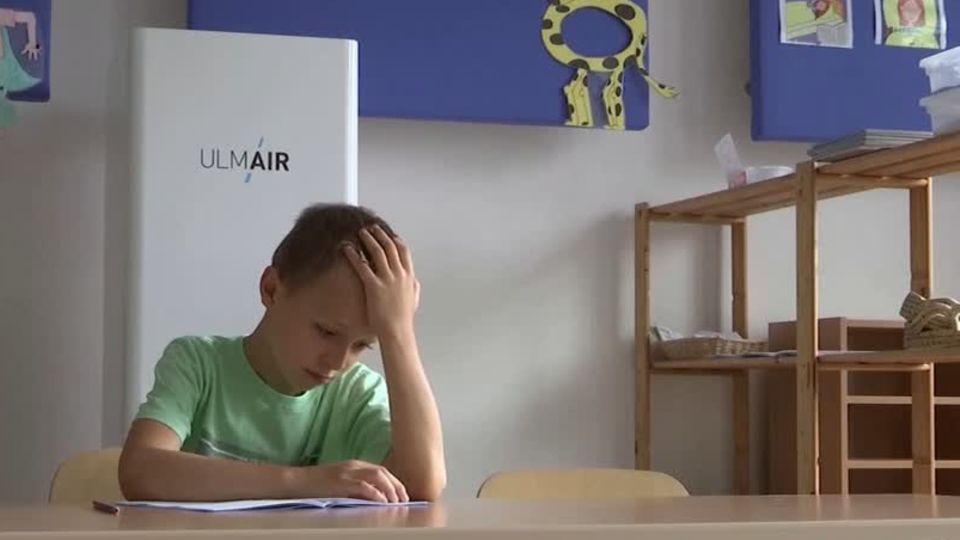 In einem Klassenzimmer sitzt ein junge in grünem T-Shirt mit aufgestütztem Kopf über einem Papier. Hinter ihm ein Luftfilter