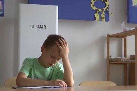 In einem Klassenzimmer sitzt ein junge in grünem T-Shirt mit aufgestütztem Kopf über einem Papier. Hinter ihm ein Luftfilter