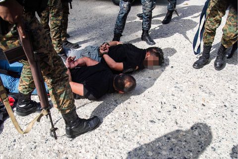 Soldaten in Tarnfleck und mit schwarzen Steifeln stehen um zwei gefesselte, am Boden liegenden Männer herum